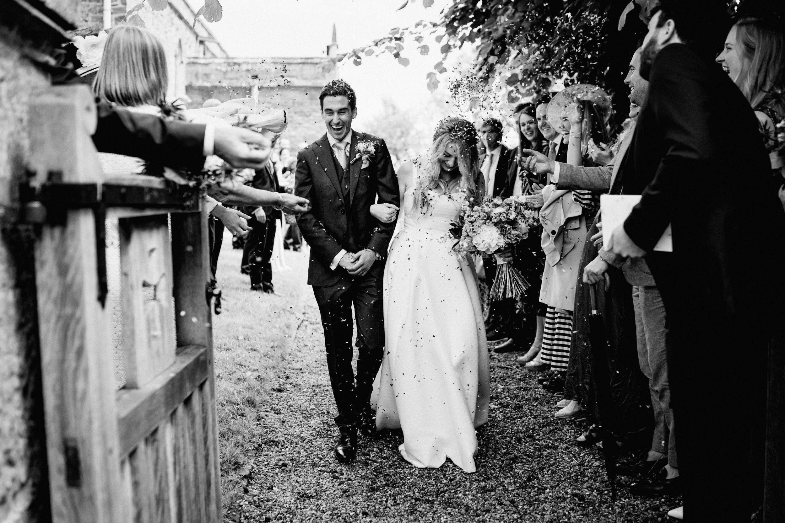 Black and white wedding photo showing newlyweds walking through guests tossing confetti in a rustic garden setting.