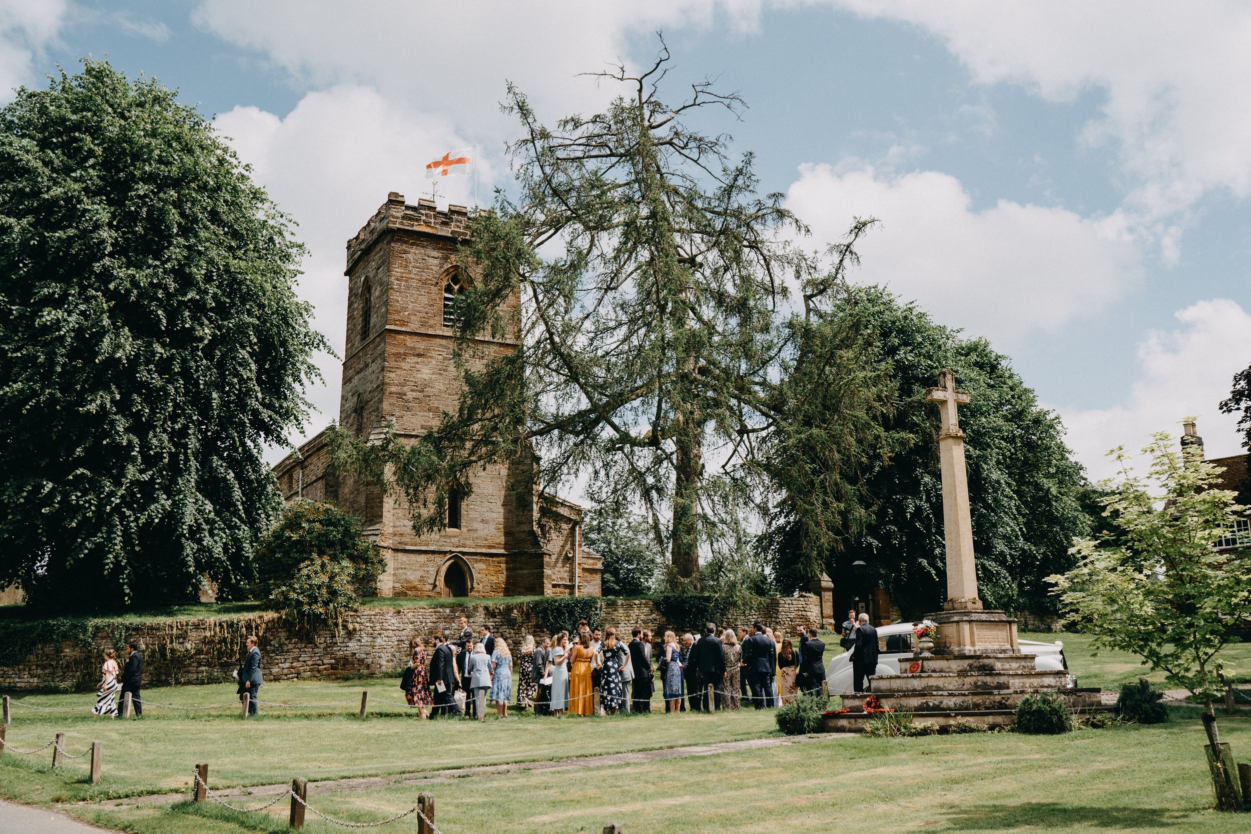 Small gathering of visitors near historic stone church tower and monument in English countryside garden setting.