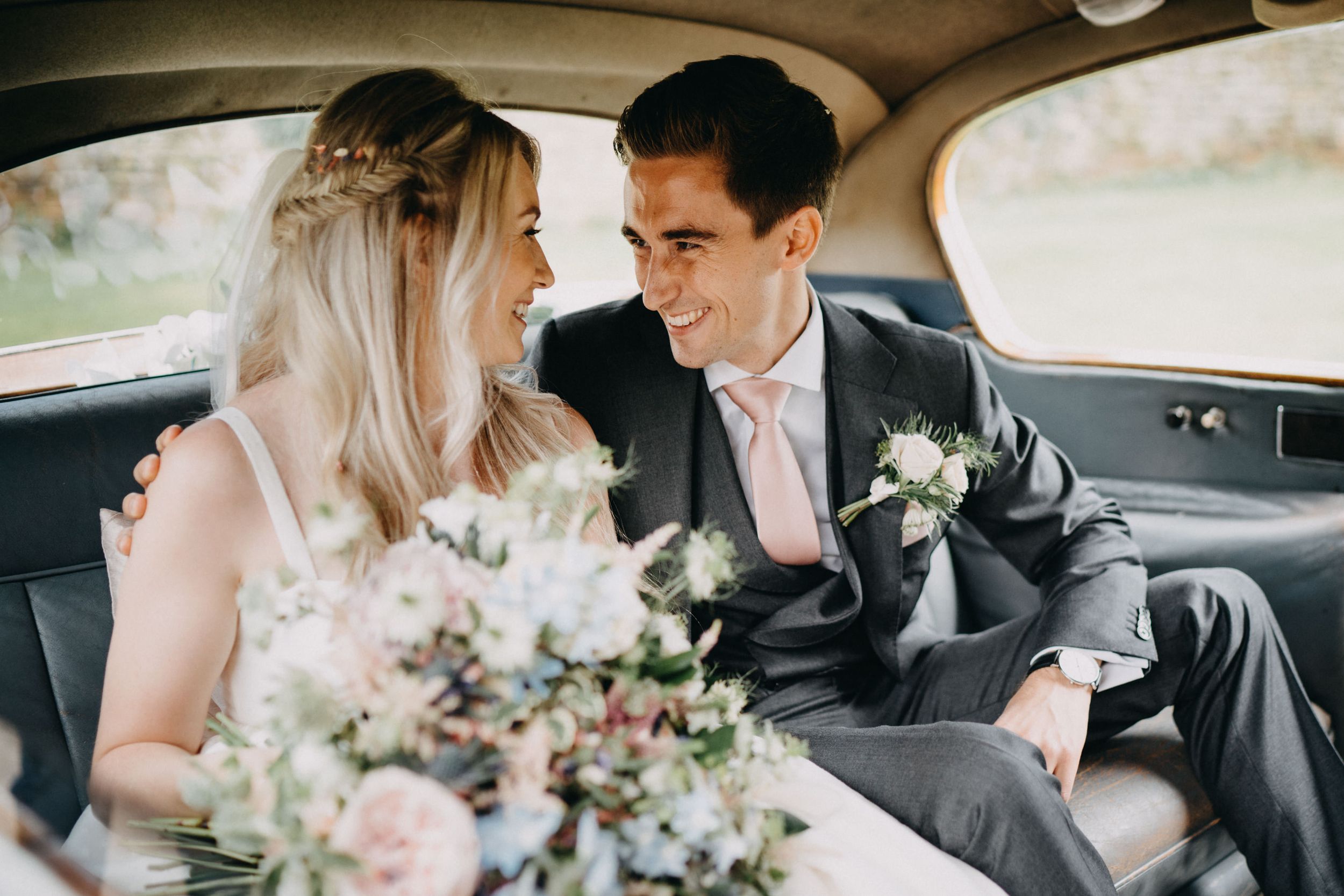 A romantic moment between newlyweds sitting in vintage car with delicate bouquet of blush pink flowers.