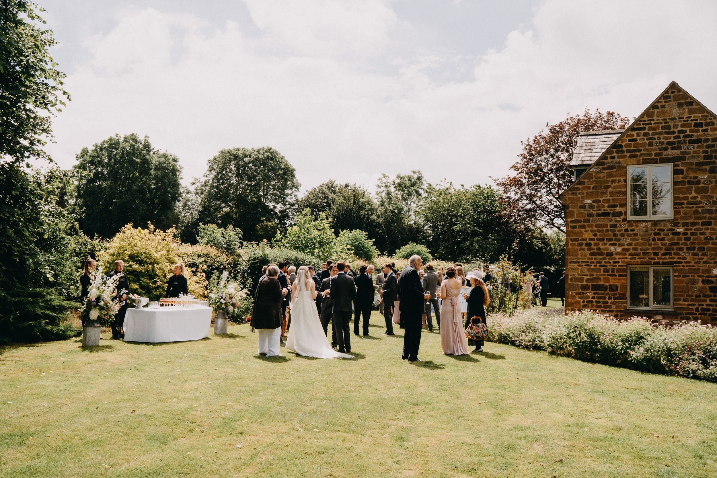Outdoor garden wedding reception with guests mingling on a summer lawn beside a rustic brick building.