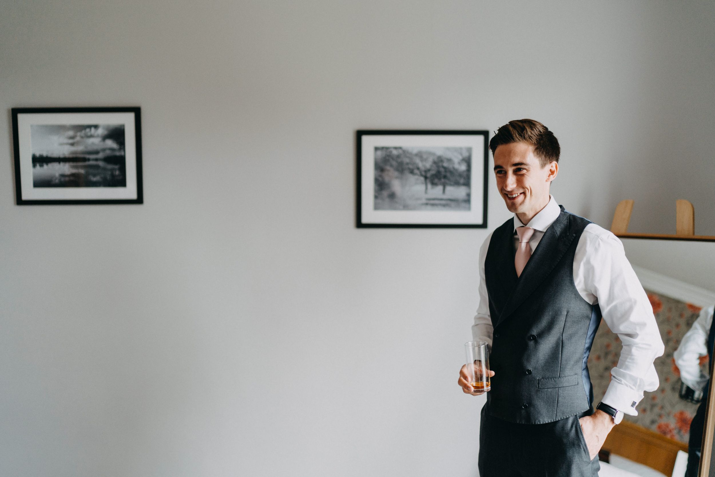 A person in a gray vest and white dress shirt holds a glass while standing by framed artwork on a wall.