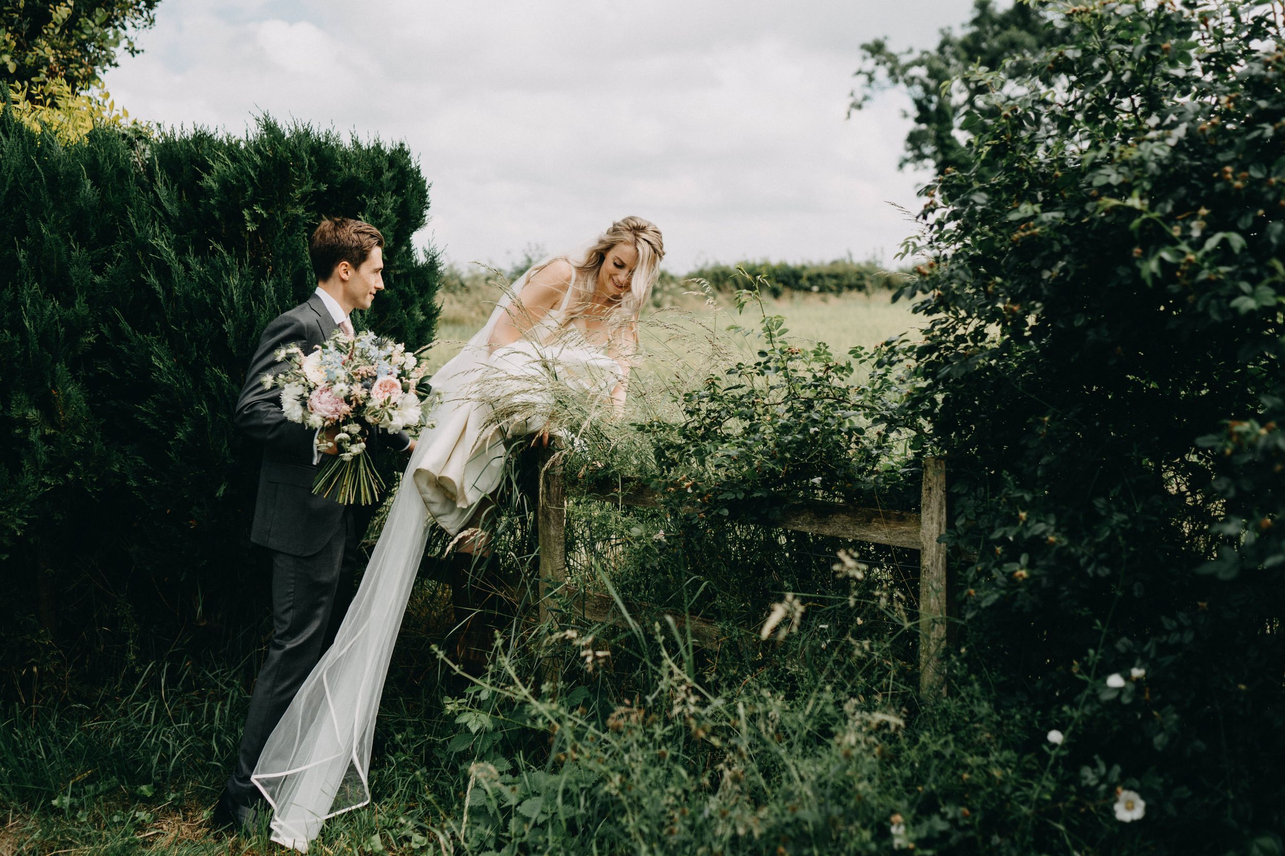 Two people in wedding attire stand among lush greenery and wildflowers on an overcast day during a countryside celebration.