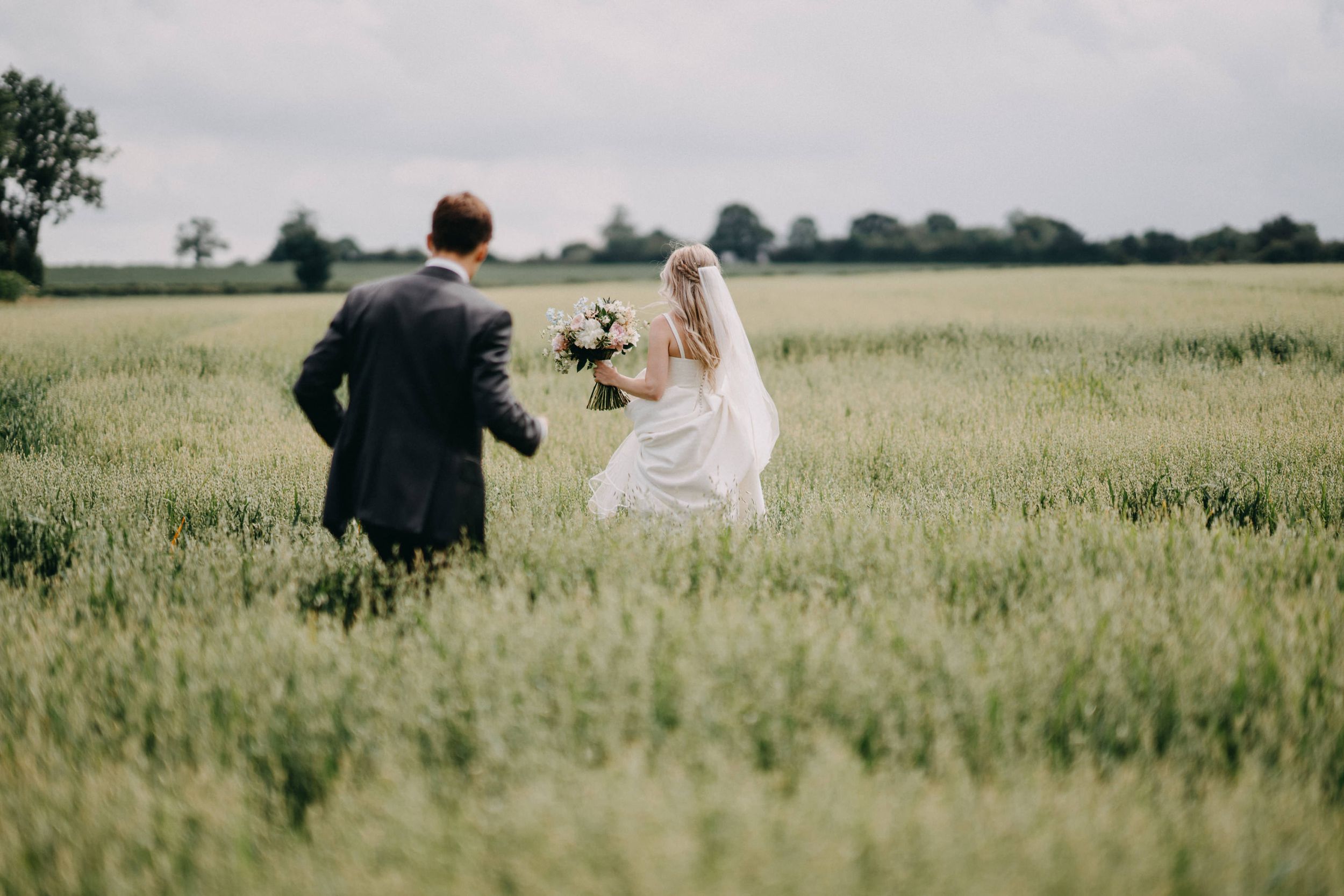 A couple in wedding attire walks through a lush green meadow under bright sunlight.