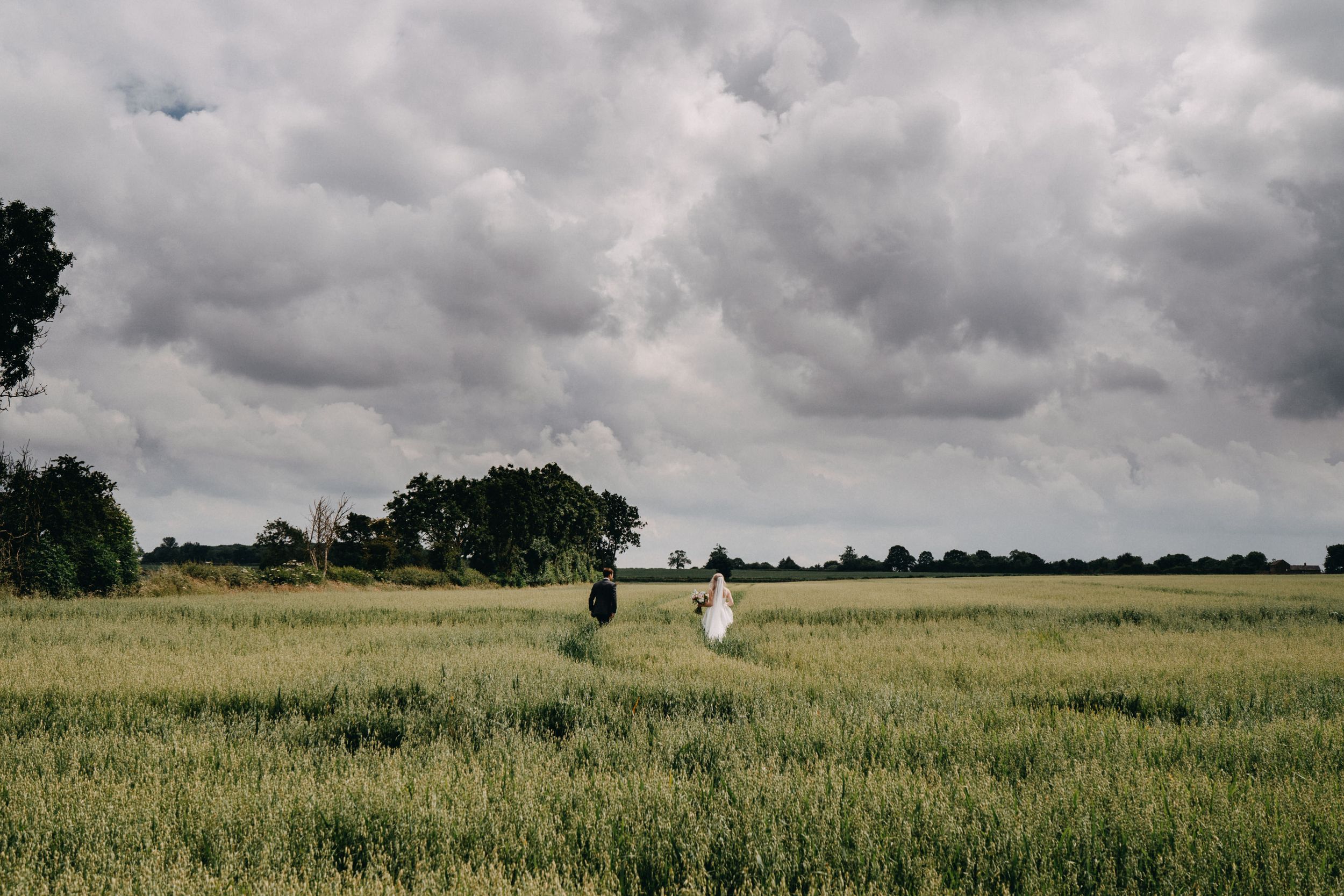 Couple stands in distance across green meadow under dramatic cloudy sky.
