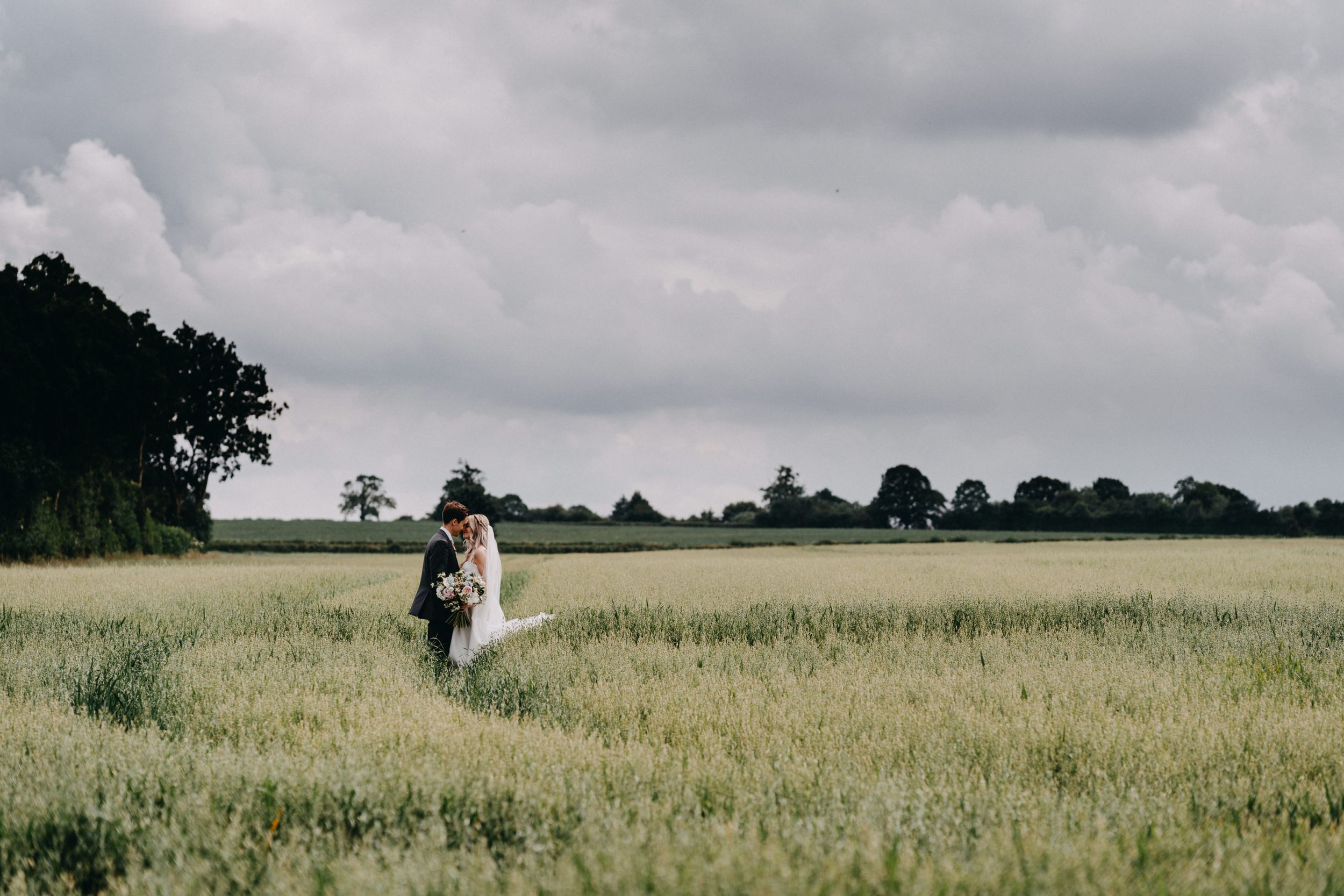 A romantic scene of a couple walking through a tall grass field under moody cloudy skies.