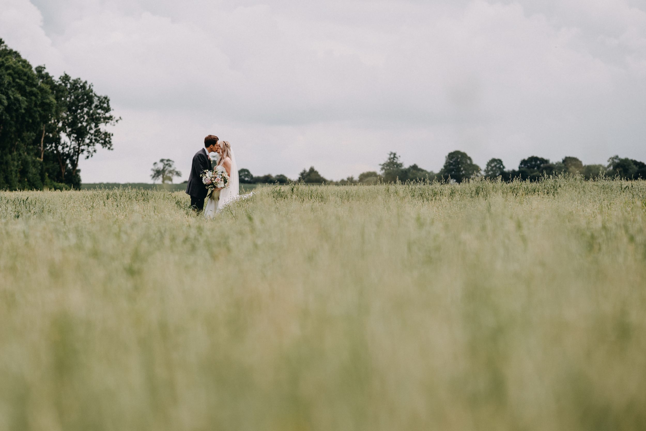 A couple stands together in a sunlit meadow of tall grass on an overcast summer day.