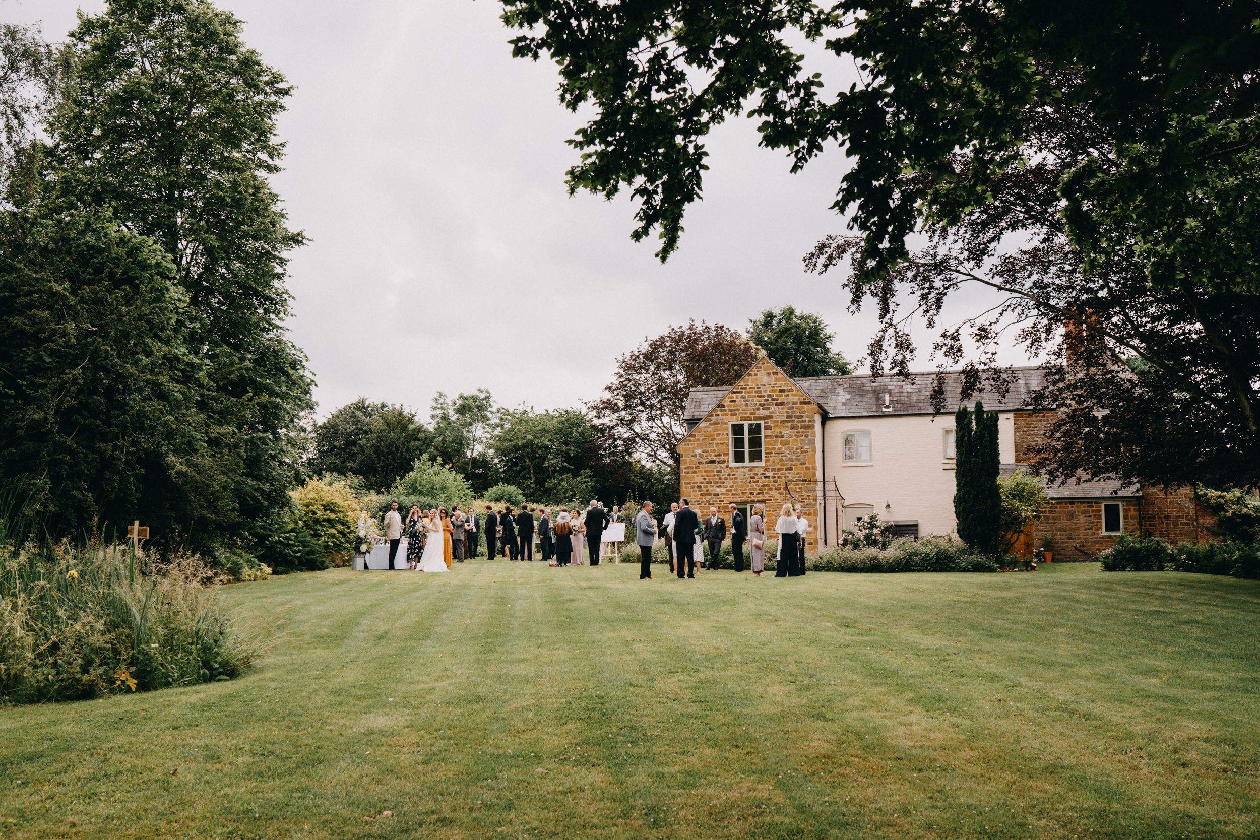 Wedding guests gather on a green lawn outside a stone country house on an overcast day.