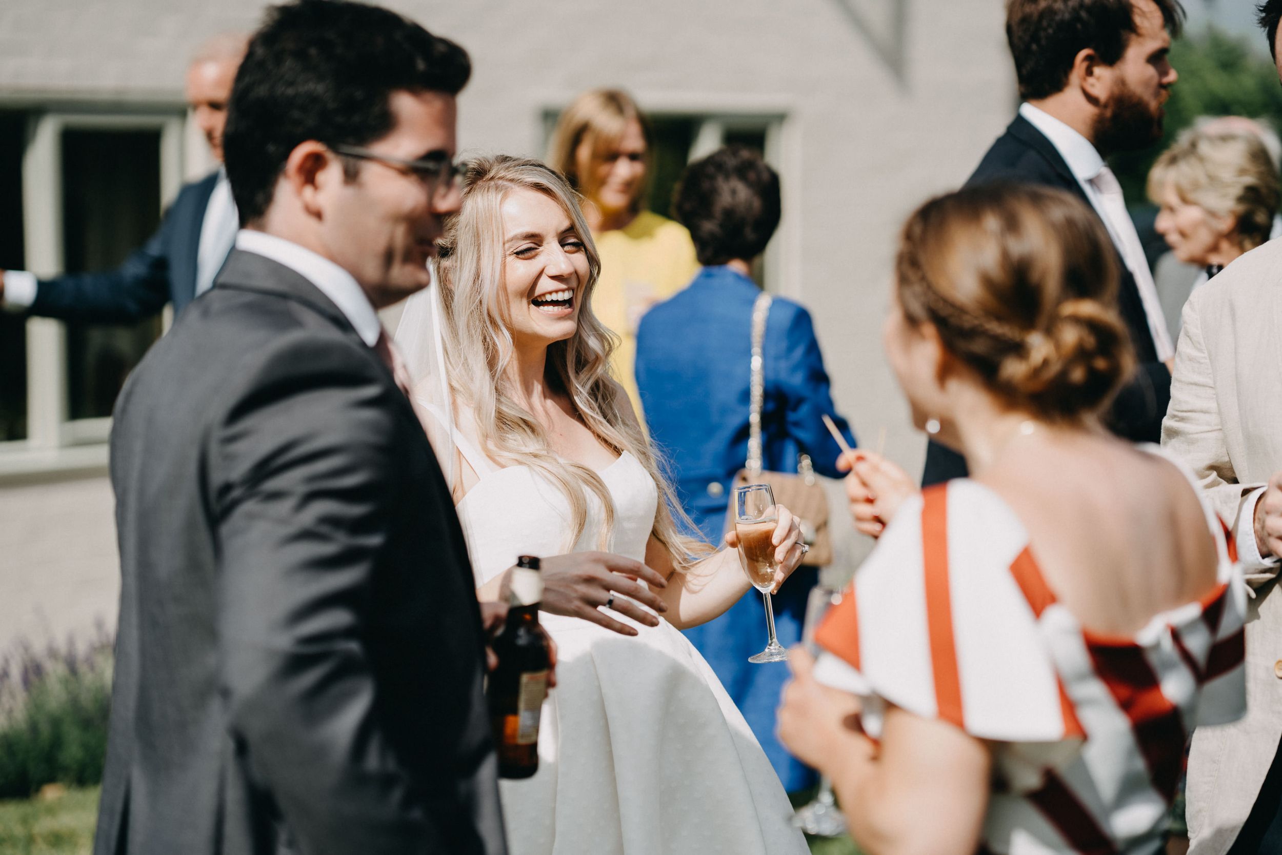 Wedding guests laugh and socialize during an outdoor reception on a sunny afternoon.