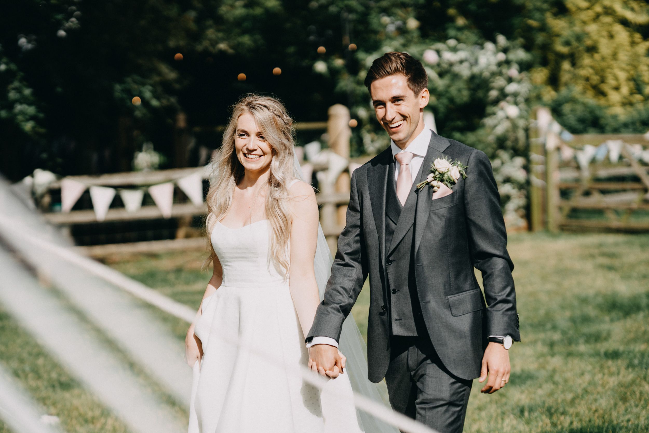 A bride and groom walk hand in hand on a sunny day with decorative bunting hanging in the background.