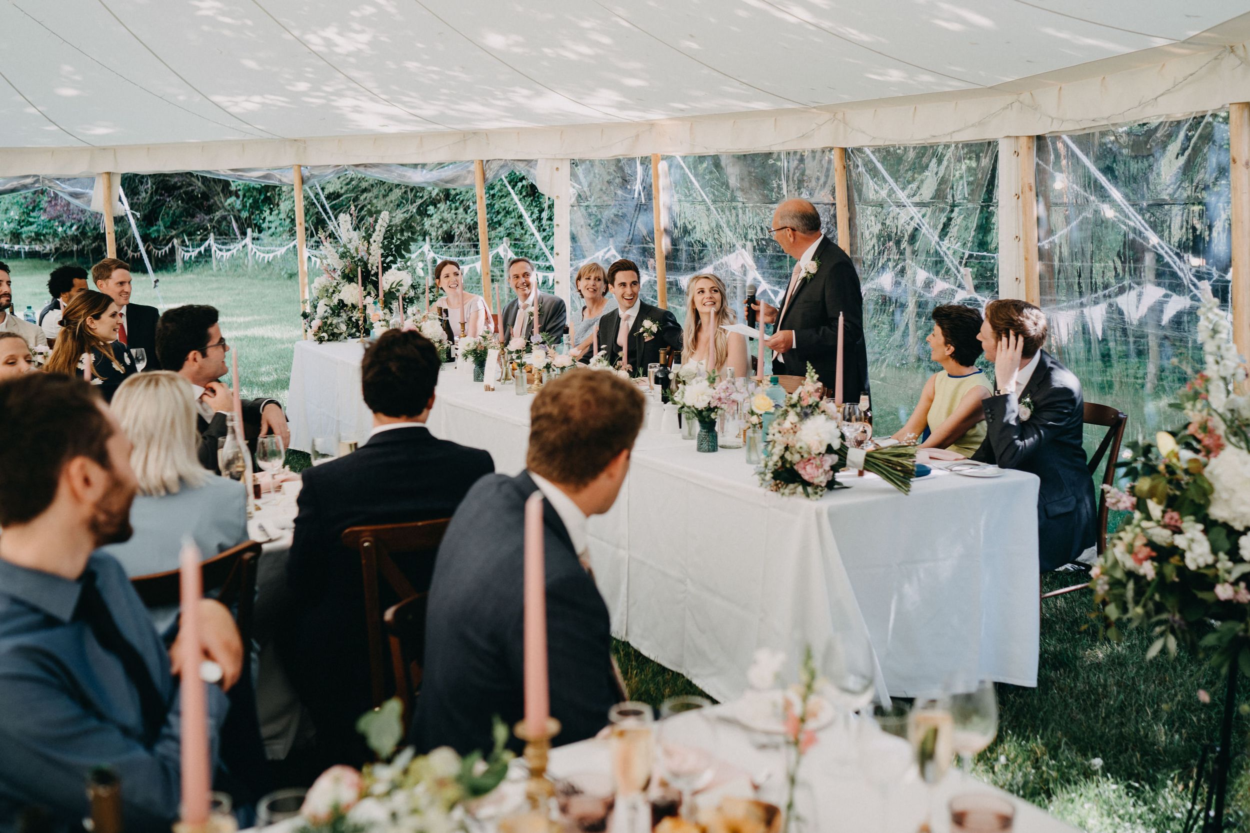 Wedding reception guests seated at tables under an elegant white tent with greenery visible through the sides.