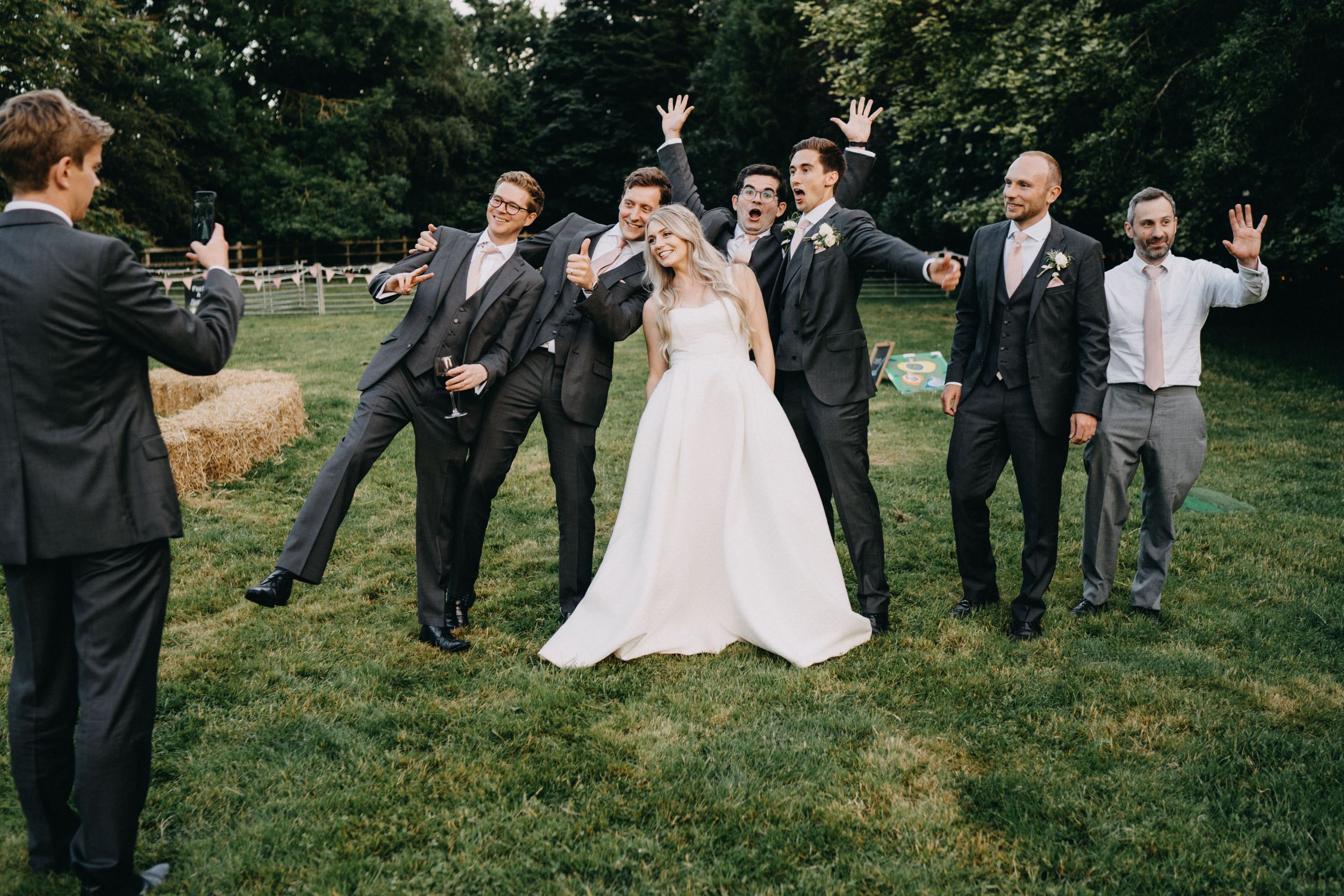Wedding party poses playfully on grassy lawn with groom aiming finger guns while others react dramatically.