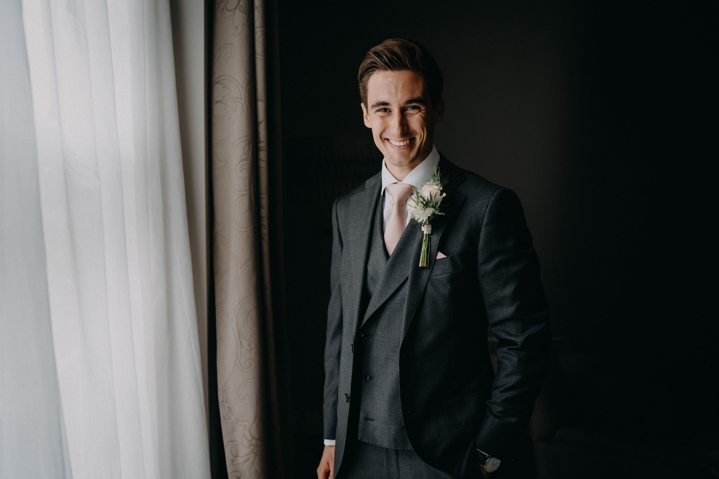 A groom in a dark gray three-piece suit stands by a window with a white boutonniere.
