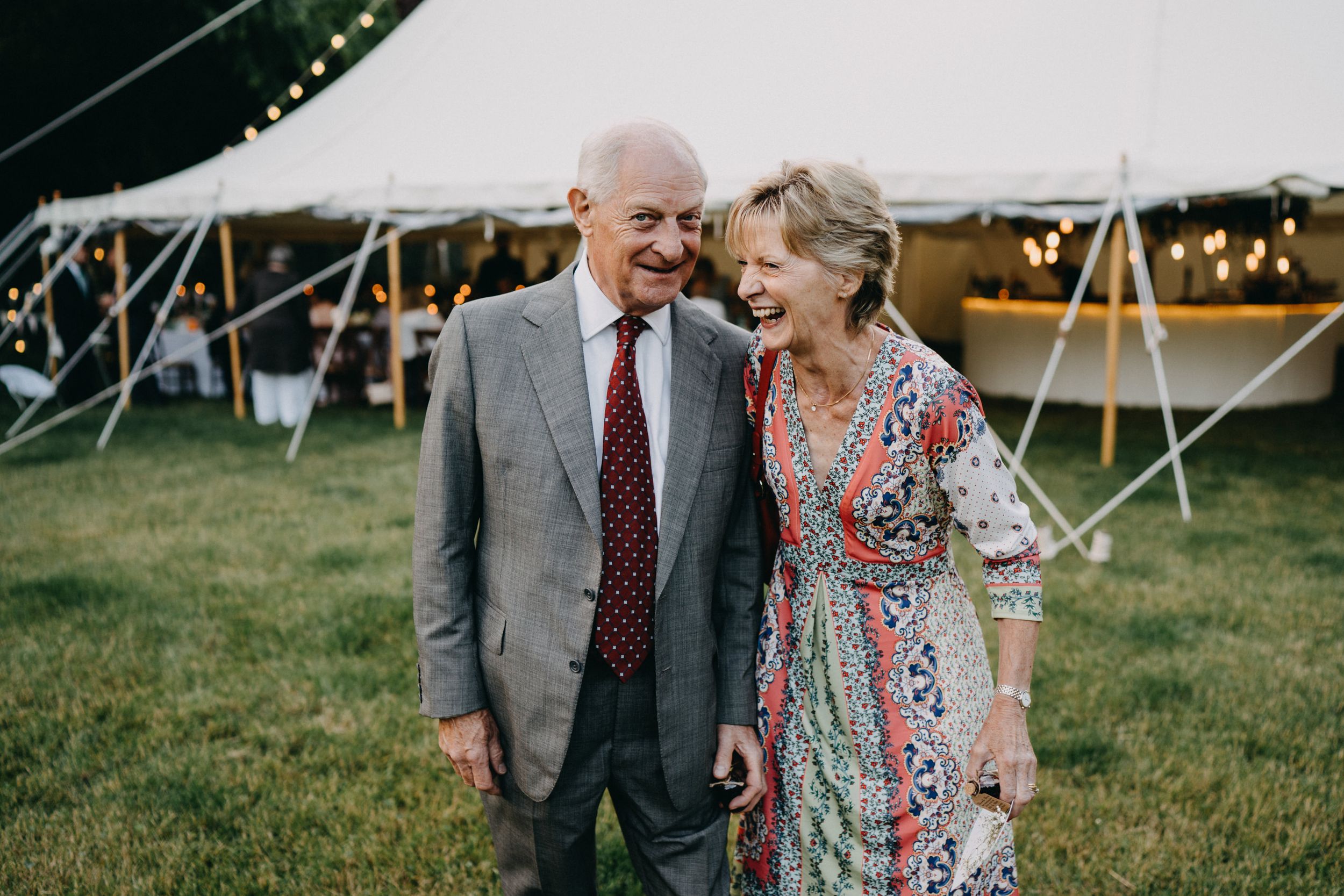 Elderly couple laughs together on grass lawn in front of reception tent with twinkling lights at outdoor event.