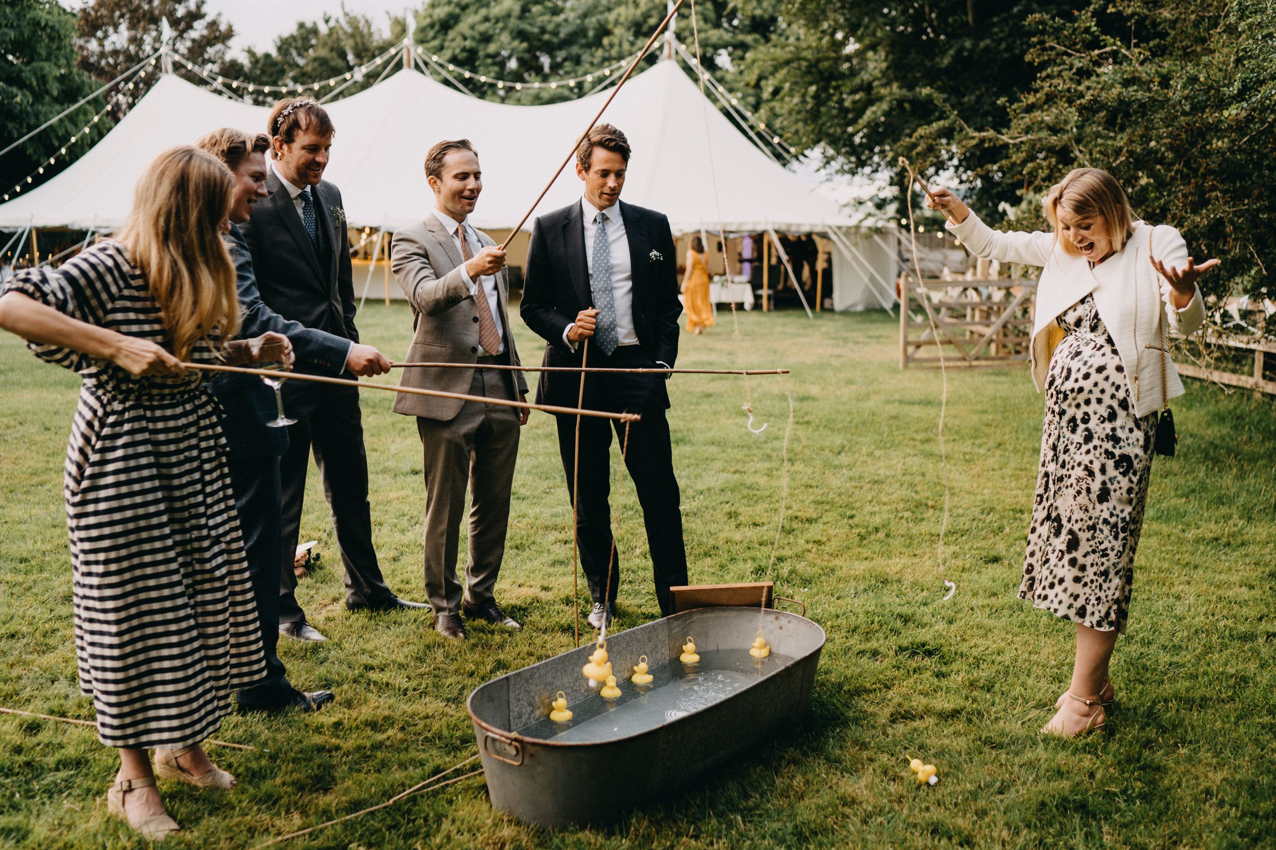 A group of wedding guests playing lawn games with a metal trough at an outdoor reception with a tent in the background.