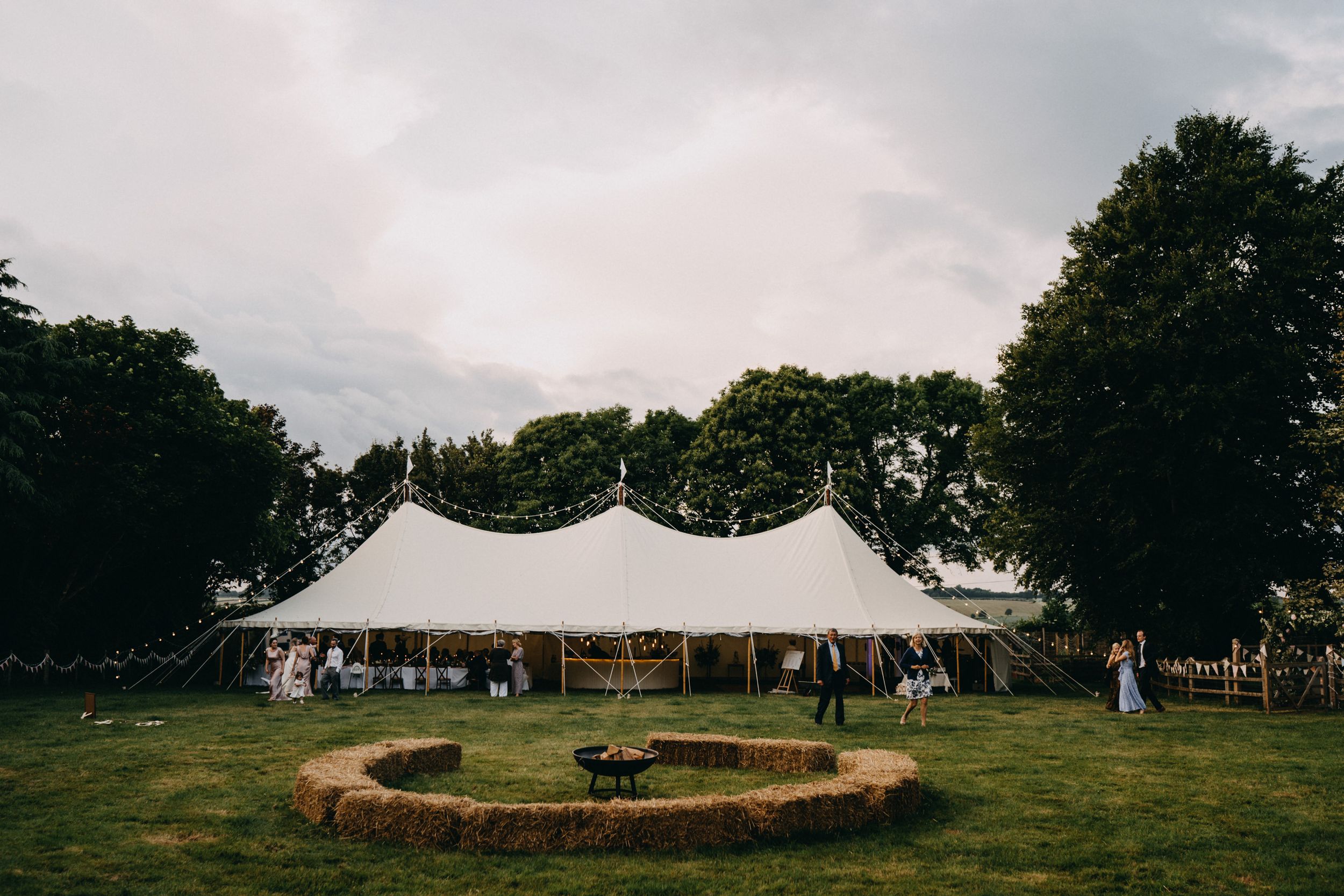 A white wedding tent stands on a grassy lawn with a circular paver patio and trees in the background at dusk.