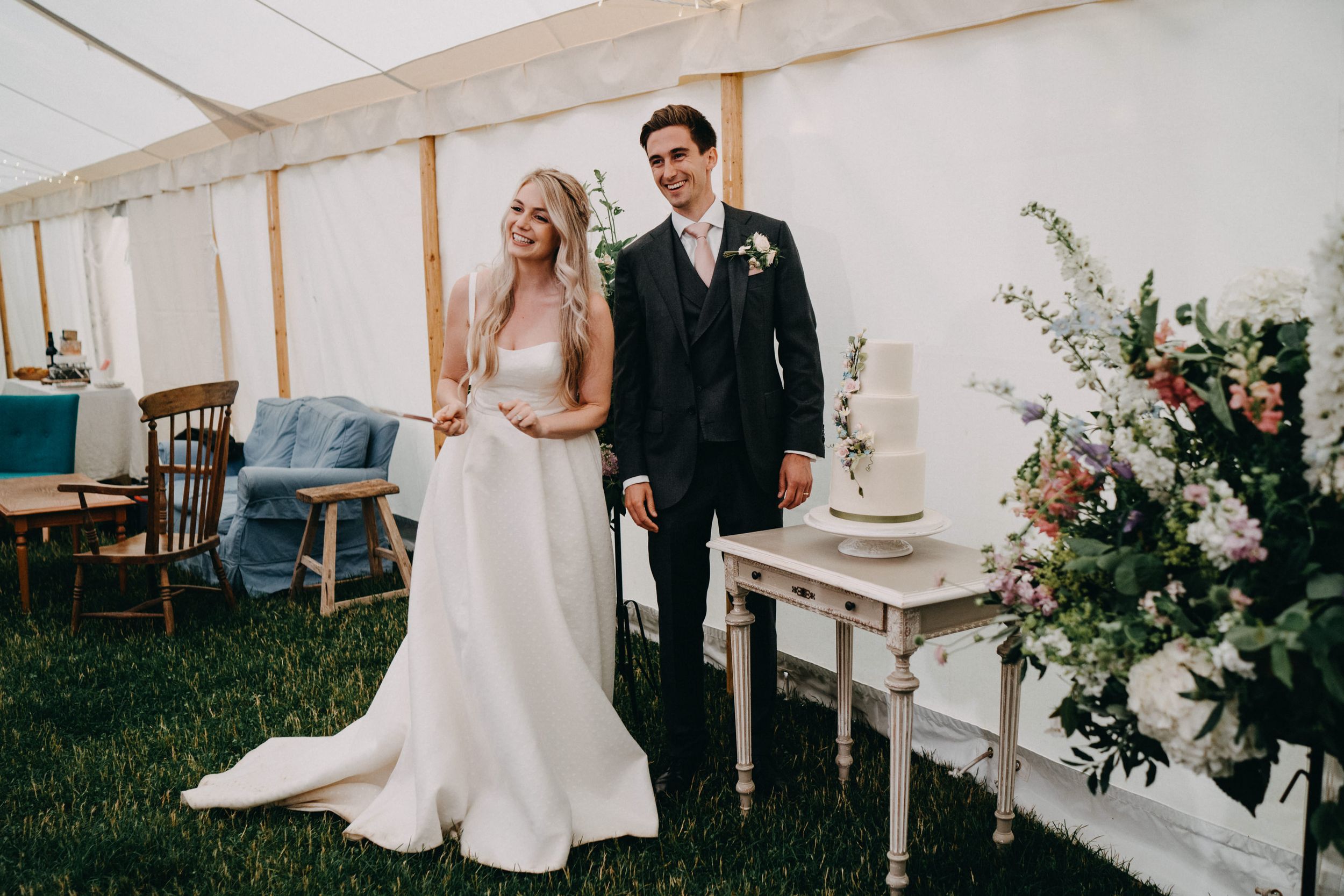 A newlywed couple stands beside a rustic white table with floral arrangements inside an elegant reception tent.