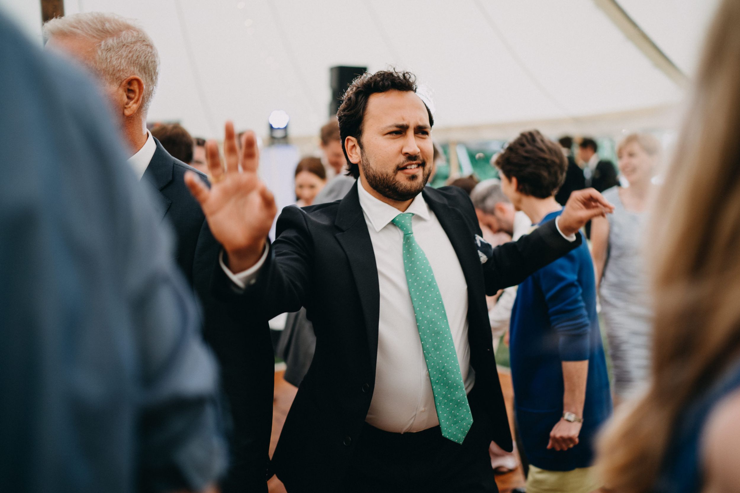 Wedding guest in black suit and mint green tie dances enthusiastically at a reception under a white tent.