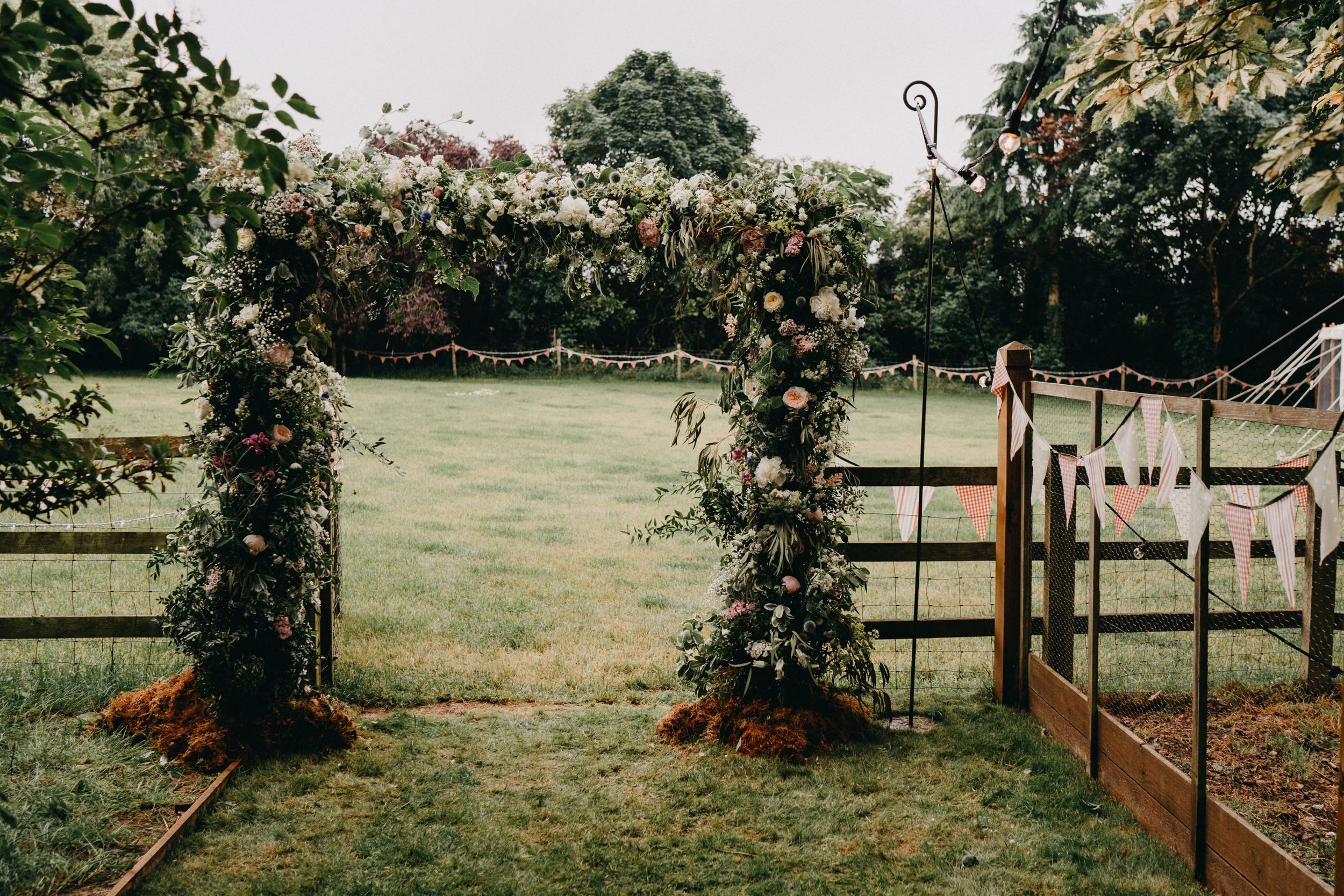 A rustic floral wedding arch with pink roses stands in a garden setting overlooking a grassy lawn.