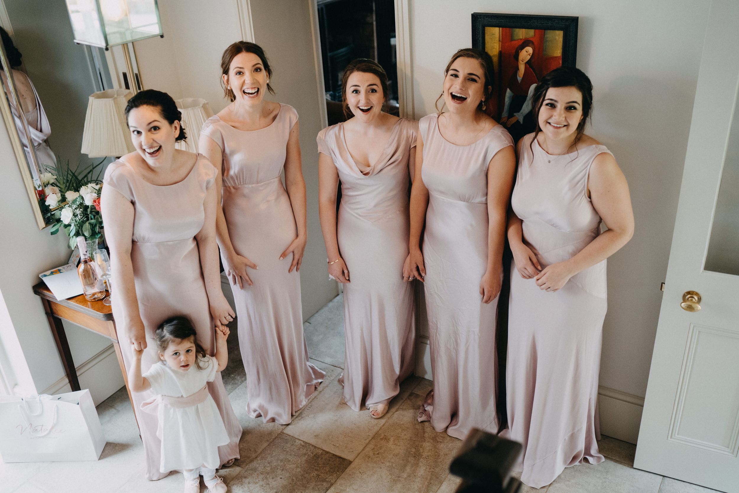 A group in matching blush pink floor-length dresses stand together with a small child in white at a formal event.