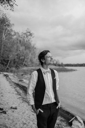 Black and white photo of someone in formal attire gazing into the distance while standing on a scenic beach shoreline.