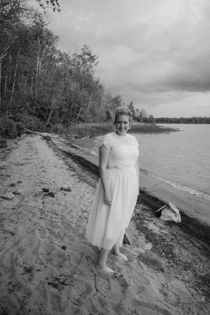 Black and white photograph of a lake shore scene with a figure in a white dress standing on a sandy beach.