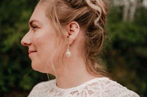 Side profile portrait showing elegant white lace dress detail and a delicate pearl drop earring against a green background.