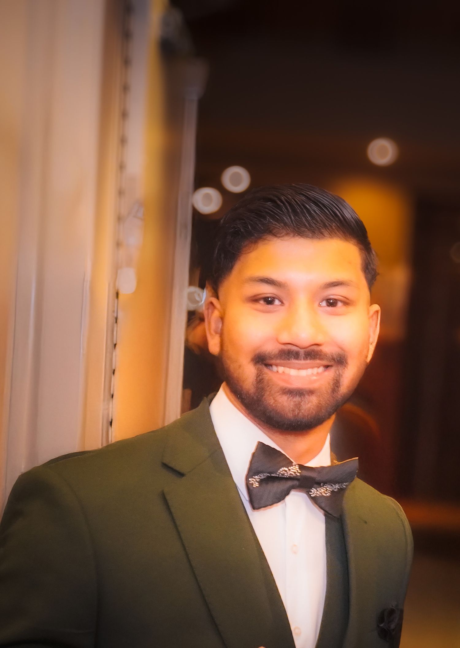 Pranay in a dark green suit and bow tie smiling during formal event, portrait with warm evening lighting