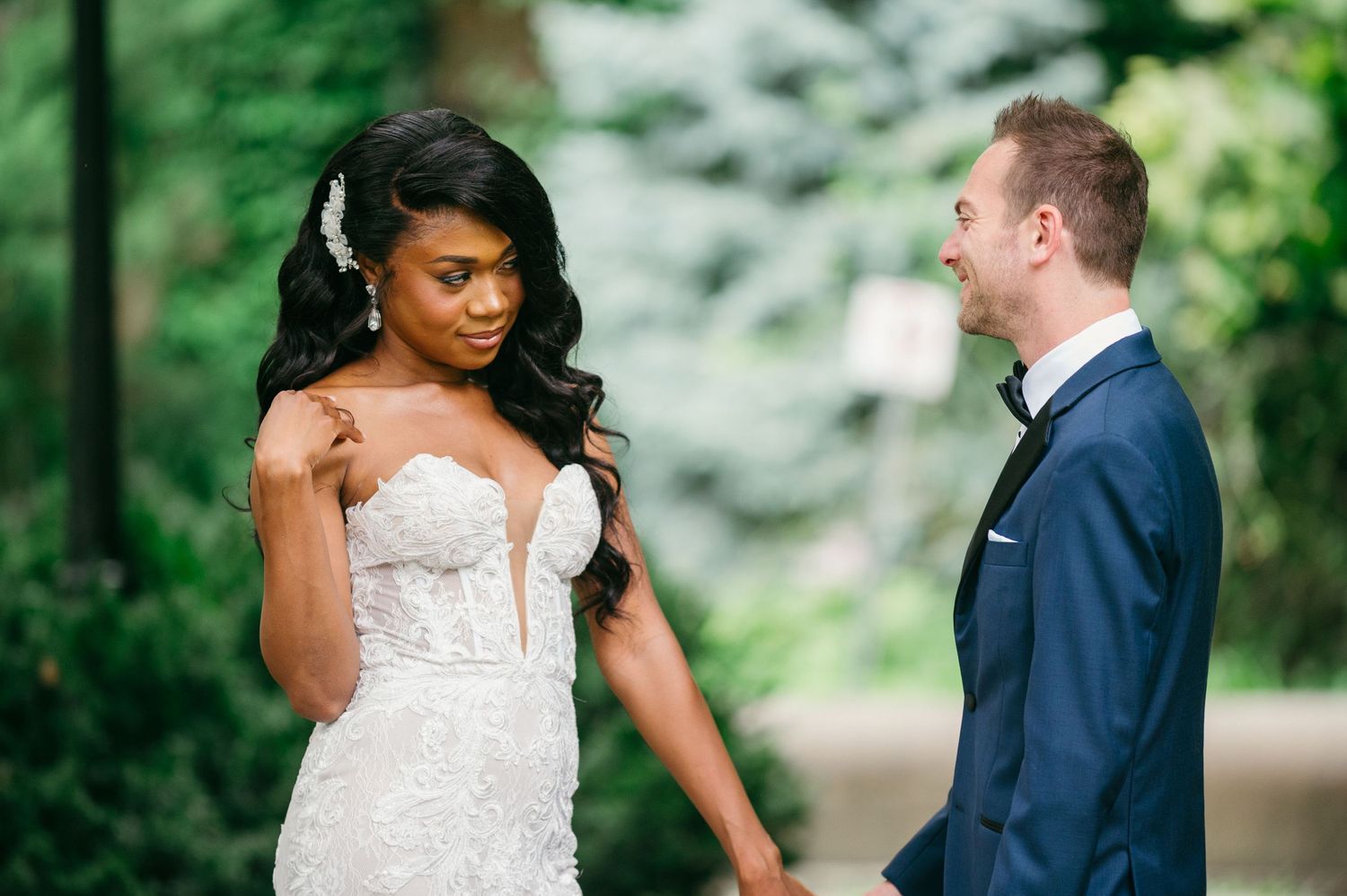 A bride in a white strapless gown and groom in navy blue suit share an intimate moment on a tree-lined path.