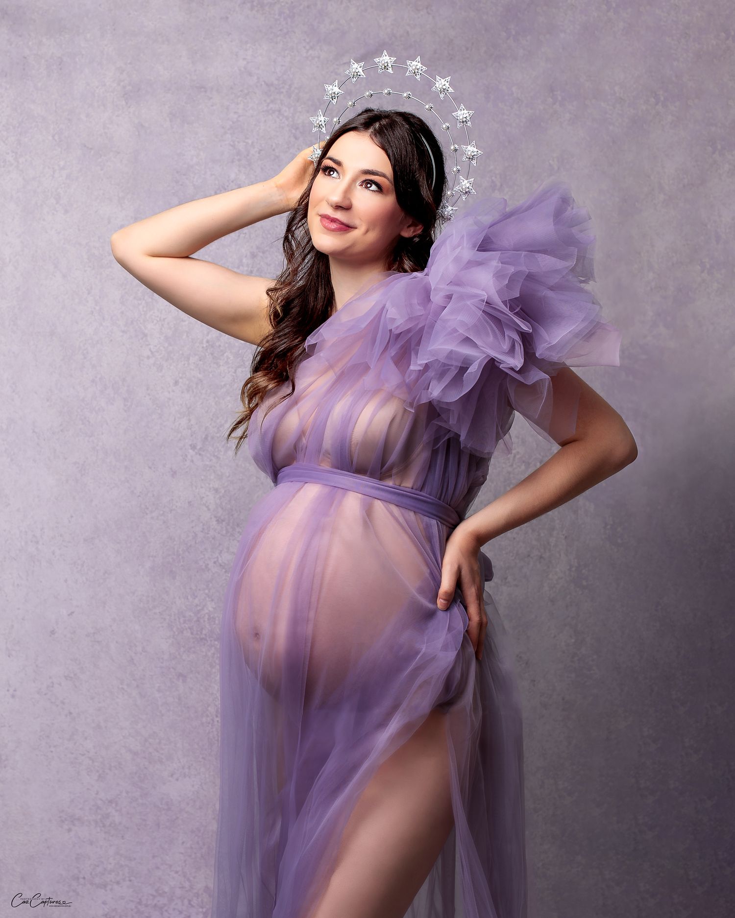 Woman in a purple gown and silver crown, arm raised above her head, graceful pose in studio.