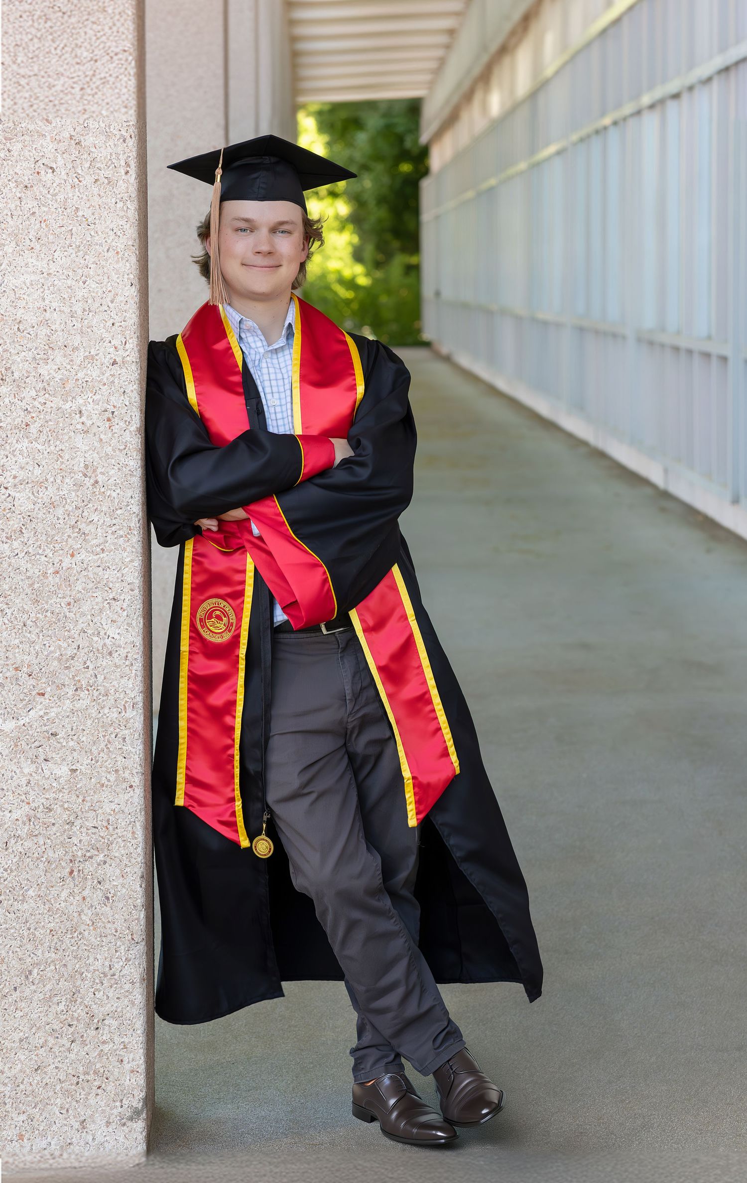 Colorado university graduate in black cap and gown with red and gold stole leans against column in hallway.