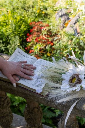 A hand rests on an open book next to a white sunflower on a rustic wooden bench in a lush garden setting.