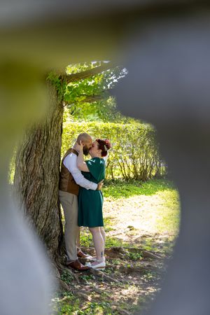 A loving couple shares a romantic embrace against a tree trunk in a sunlit woodland setting.