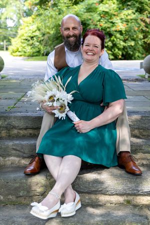 A couple in matching emerald green outfits sit together on stone steps holding white flowers.