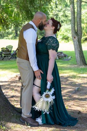 A couple in formal attire share an intimate moment on a log in a sunlit forest setting, she in emerald green, he in brown.