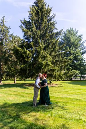 A couple embraces on a lush green lawn with tall evergreen trees creating a scenic backdrop on a sunny day.