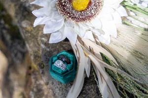 A bright teal engagement ring nestles among white daisy petals in a rustic outdoor setting.