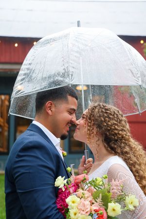 A romantic couple shares a kiss under a clear umbrella while holding colorful wedding bouquets.