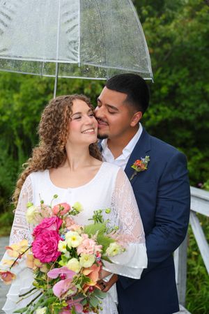 A tender moment shared between a couple on their wedding day under transparent umbrellas in a lush garden setting.