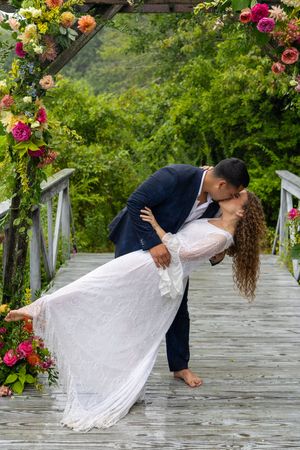 A couple in wedding attire perform a romantic dip pose on a rustic wooden walkway surrounded by lush greenery and flowers.