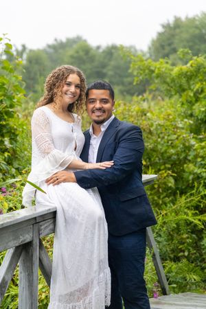 A couple in wedding attire embrace romantically on a wooden deck surrounded by lush greenery on a summer day.