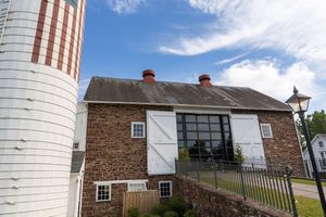 Historic stone barn building with white trim and large loading doors sits next to a tall white silo under blue skies.