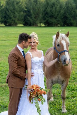 A couple in wedding attire stands with a palomino horse in a green field with trees in the background.