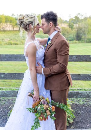 A lush rustic outdoor wedding scene shows newlyweds embracing in front of a black fence with autumn florals.