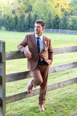 A person in a brown suit leans casually against a wooden fence in a pastoral countryside setting with green trees.