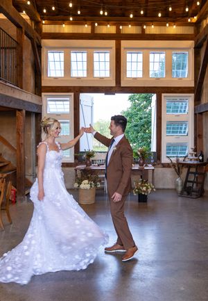 Wedding couple dances together in rustic barn venue with string lights overhead during first dance celebration.
