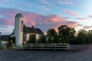 A rustic farm with white silo and barn stands against a dramatic pink and orange sunset sky.