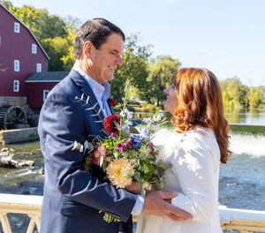 A couple shares a romantic moment by a rustic red barn and pond on a sunny autumn day.