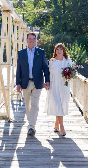 A couple walks hand in hand across a sunlit wooden bridge wearing white and navy blue wedding attire.