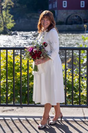 A person in a white dress holds a burgundy and pink floral bouquet while standing on a wooden deck near water.
