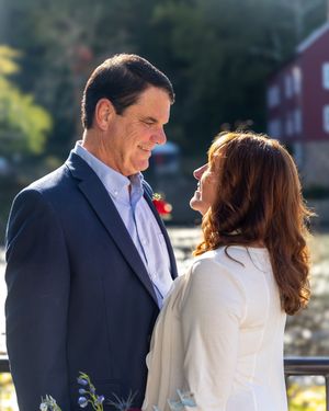 A couple embraces on an outdoor deck with a rustic red building visible behind them.